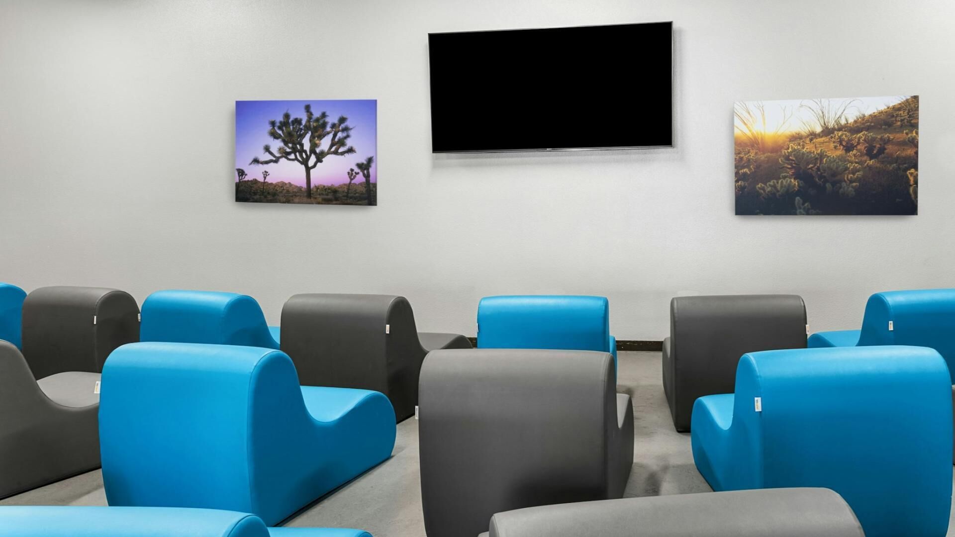 Une pièce avec des fauteuils rembourrés aux couleurs bleu et gris, disposés en rangées, un téléviseur à écran plat fixé au mur et deux tableaux représentant des paysages, encadrés.