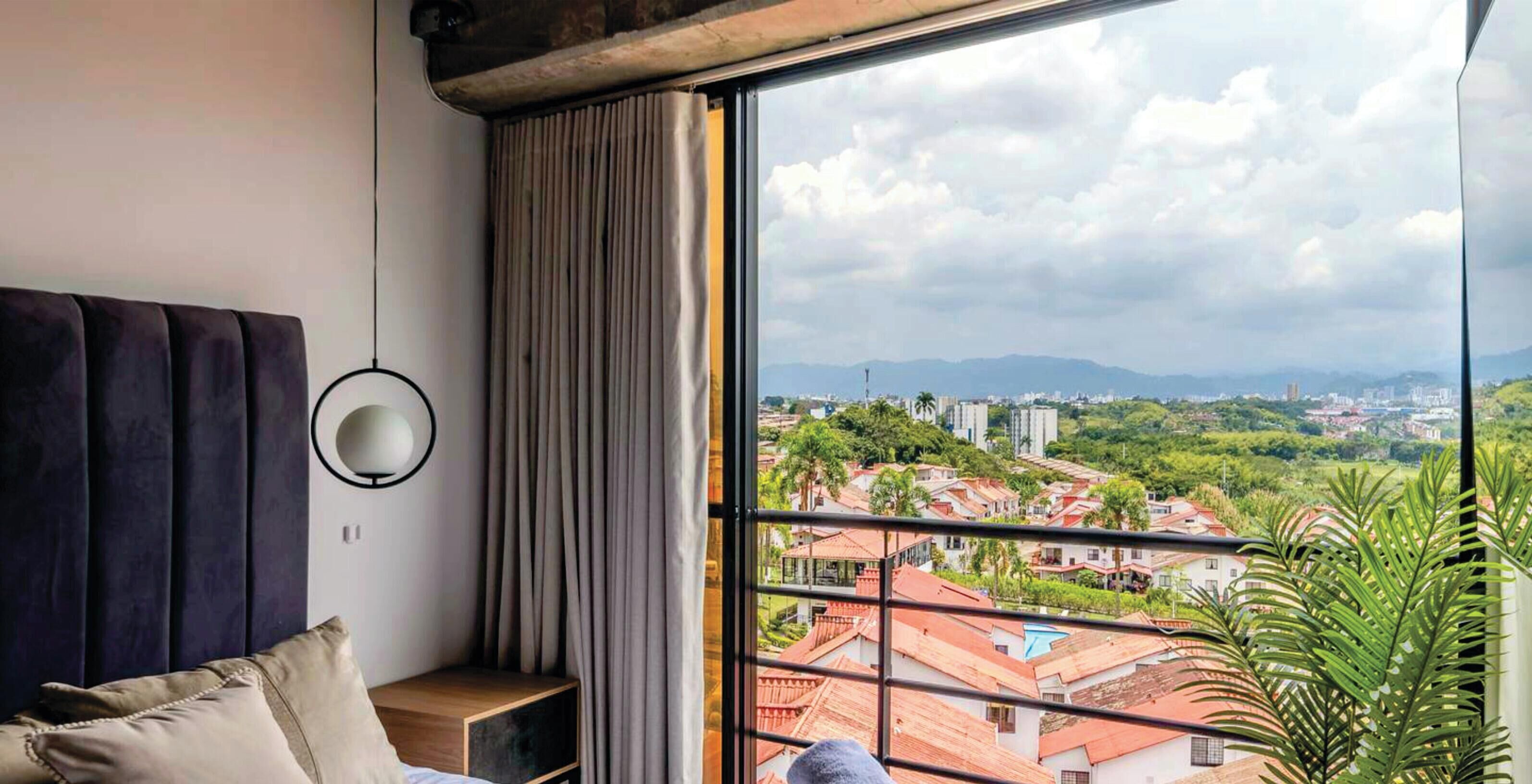 A bedroom with a large window, a bed, a bedside table, and a view of a cityscape.