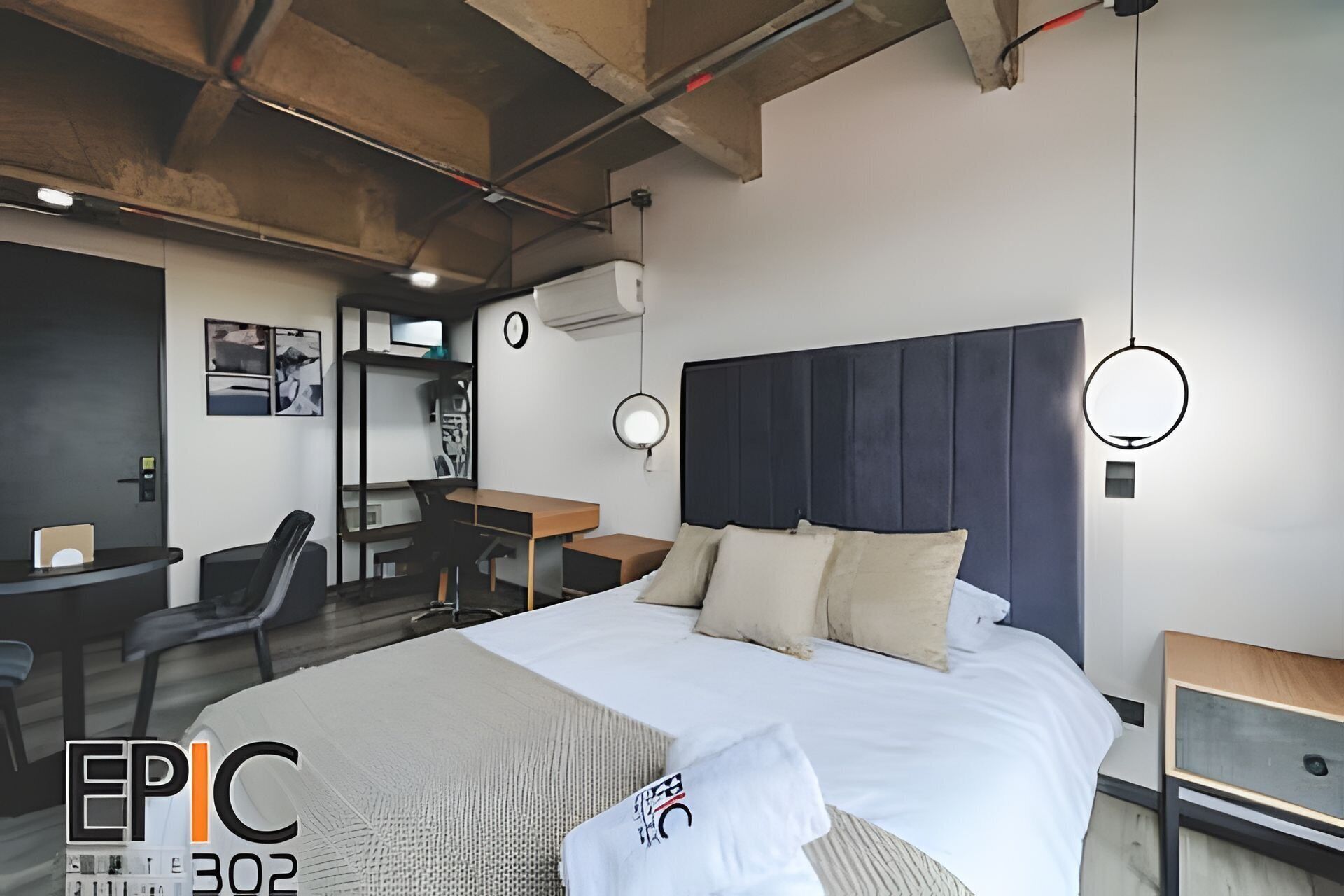 Une chambre d’hôtel moderne avec un grand lit, un bureau et une chaise. Il y a une horloge au mur et un luminaire rond au-dessus du lit.