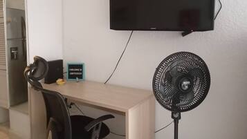 A room with a desk, a black chair, a standing fan, a mounted TV, and a refrigerator.