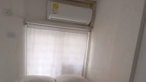 A small, single-bed room with a plain white bedspread, a window with a sheer curtain, and a wall-mounted air conditioning unit.