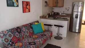 A small living area with a colorful sofa, a kitchen with a stainless steel refrigerator, and a bar stool.