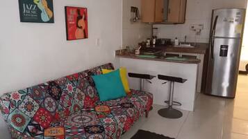 A small living area with a colorful sofa, a kitchen with a stainless steel refrigerator, and a bar stool.