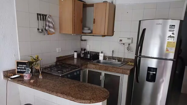 A kitchen with a stainless steel refrigerator, a stove, and a sink. There are two black bar stools and a sign on the counter.