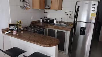A kitchen with a stainless steel refrigerator, granite countertops, wooden cabinets, a gas stove, and two black bar stools.