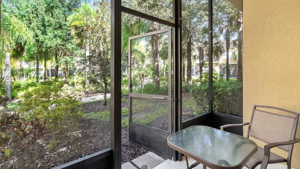 Un balcon vitré, agrémenté d’une table et de chaises, qui offre une vue sur un jardin luxuriant, où poussent différentes plantes et arbres.