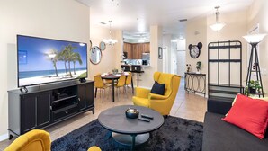 Un salon comprenant un téléviseur, un fauteuil jaune, une table à manger, un coin cuisine et un escalier.