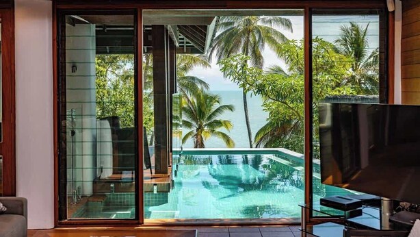 Une chambre moderne dotée d’une porte vitrée donnant accès à une piscine privée, entourée d’une végétation luxuriante et offrant une vue sur la mer.