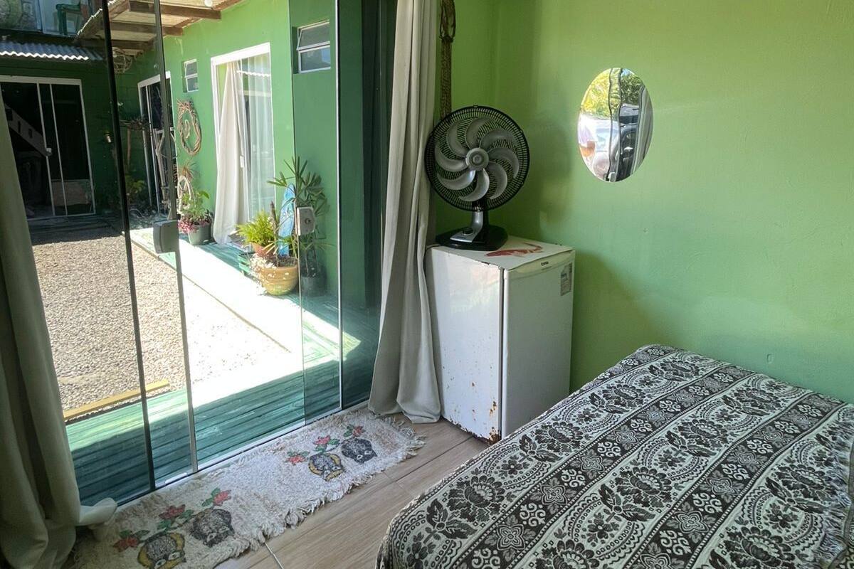 A room with a bed, a small white refrigerator, a fan, and a sliding glass door leading to an outdoor area with plants.