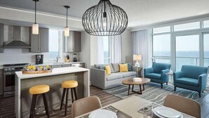 Moderni hotellihuone, jossa on keittiö, oleskelutila sohvineen ja nojatuoleineen, ruokailutila pöytineen ja tuoleineen sekä suuret ikkunat, joista on näkymä.