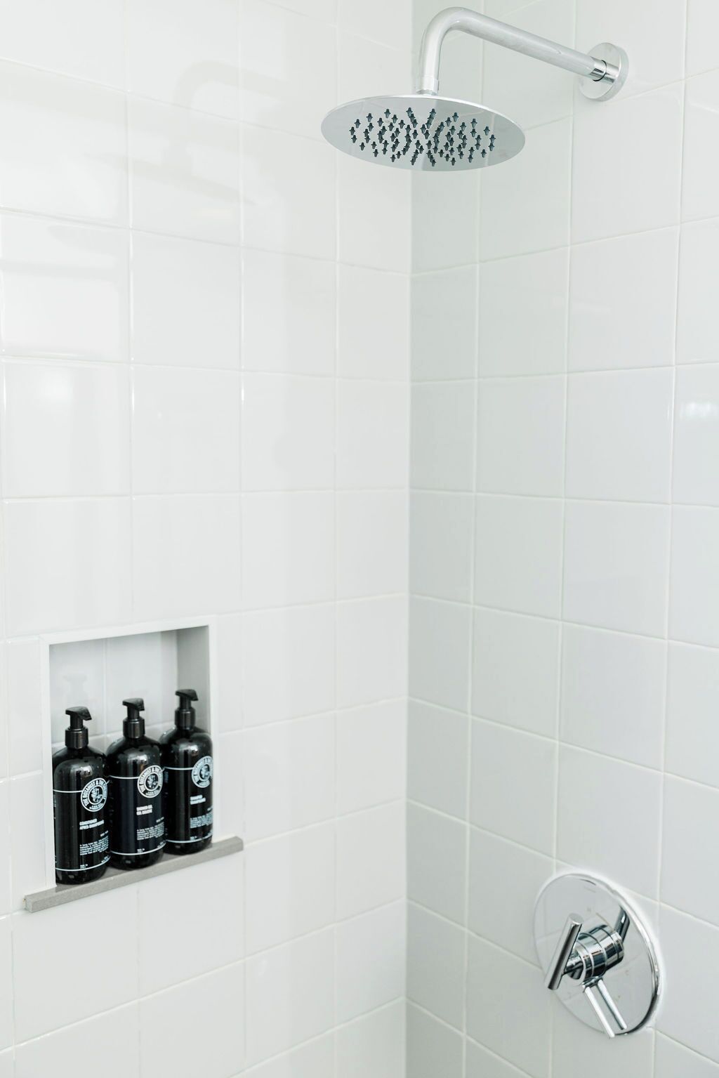 Un baño con paredes de azulejos blancos, un cabezal de ducha, un estante empotrado con botellas negras y un grifo cromado.