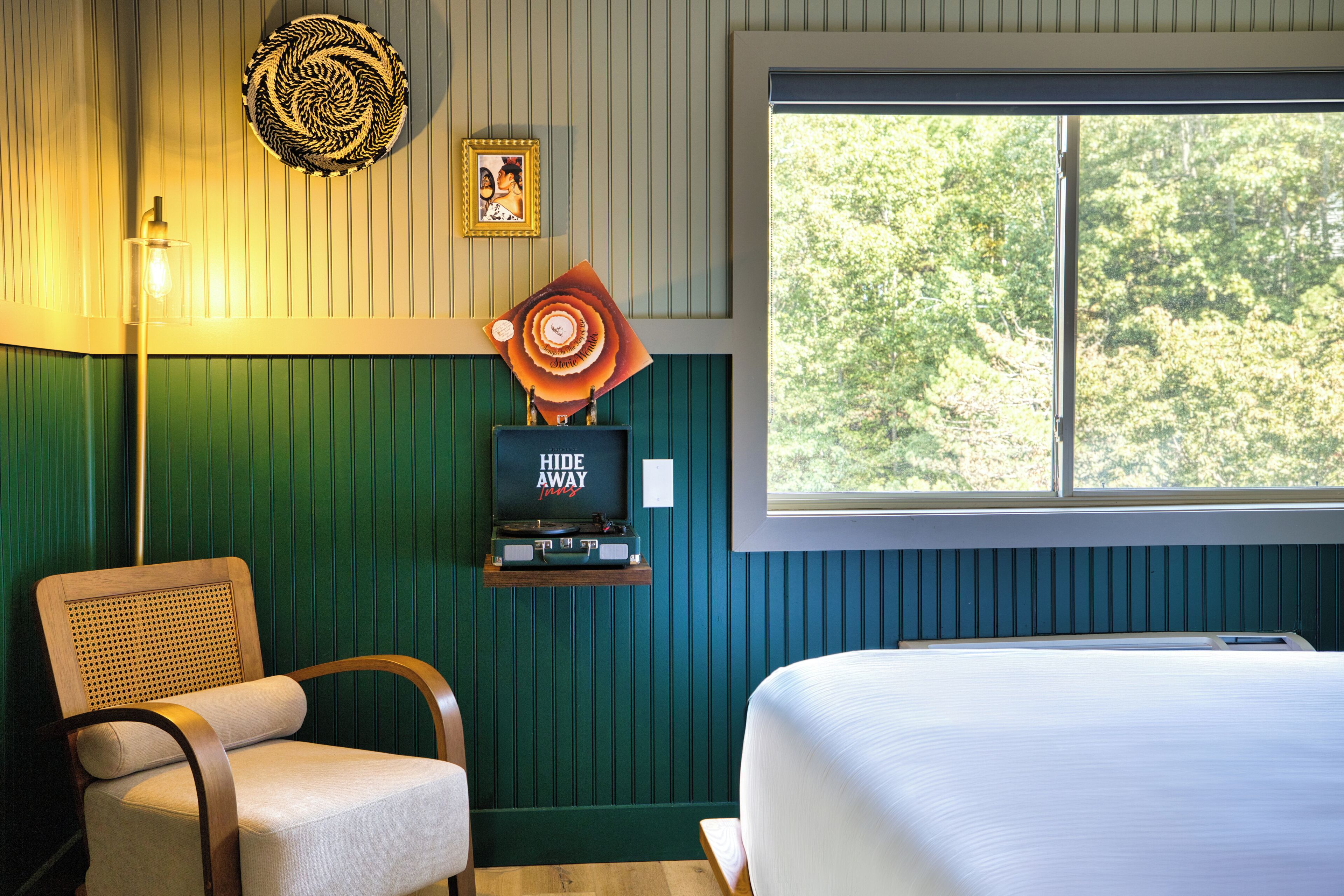 Habitación con cama, una silla, televisor montado en la pared y ventana con vista a árboles.