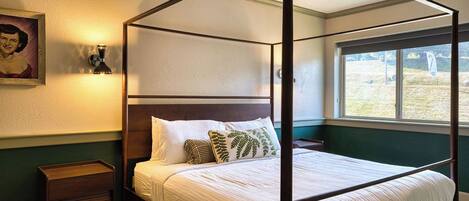 Un lit à baldaquin avec du linge de lit blanc et des coussins décoratifs, une table de chevet en bois, une lampe fixée au mur et une fenêtre donnant sur l’extérieur.