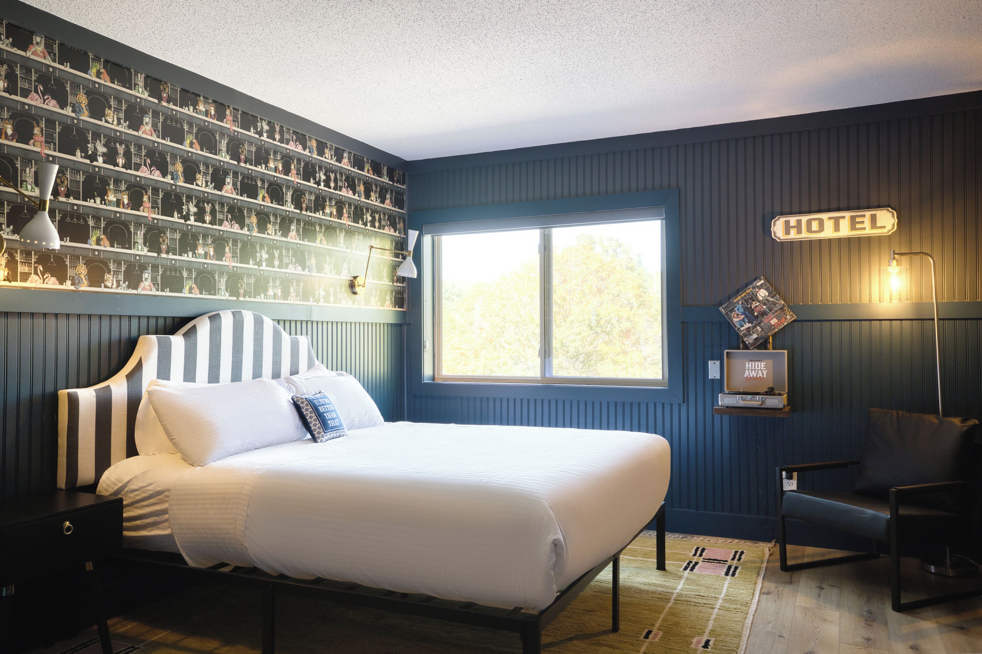 Una habitación de hotel con cama, una silla, un ventanal grande, una pared con el cartel 'HOTEL' y un estante con varios objetos.