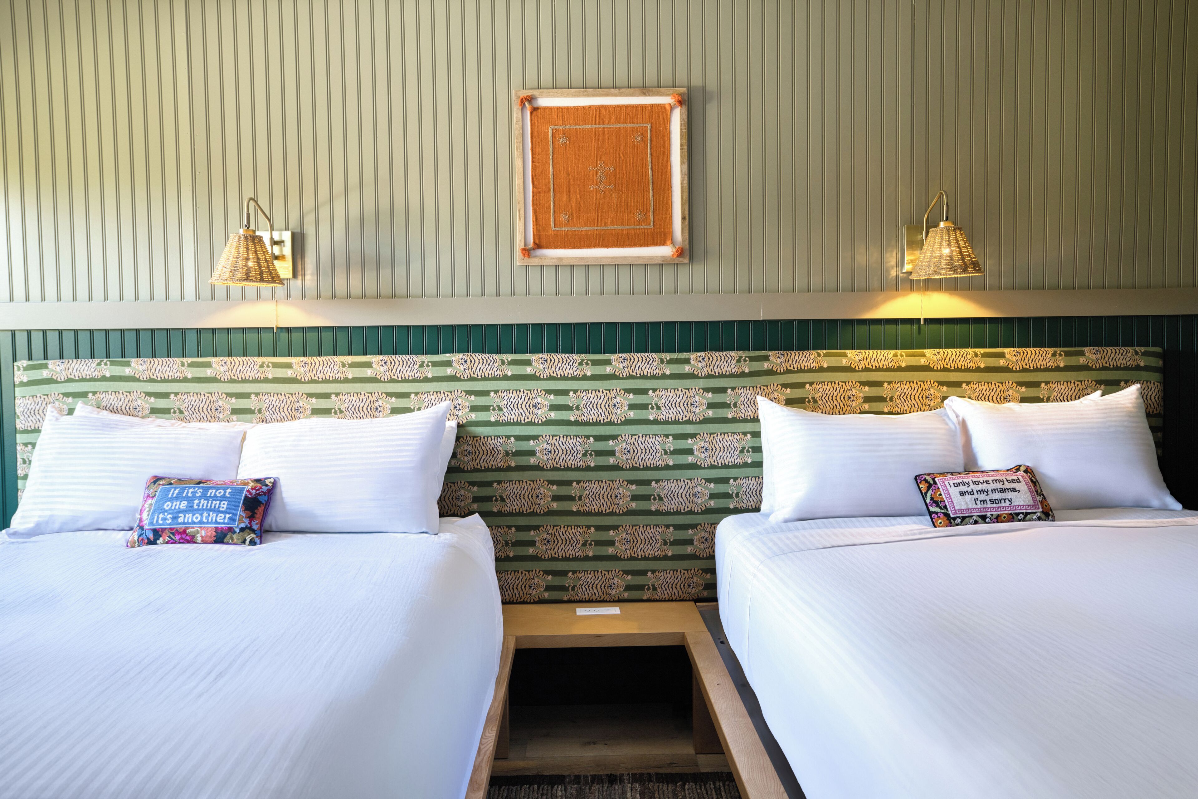 A hotel room with two beds, a patterned headboard, wall-mounted lamps, and decorative pillows.