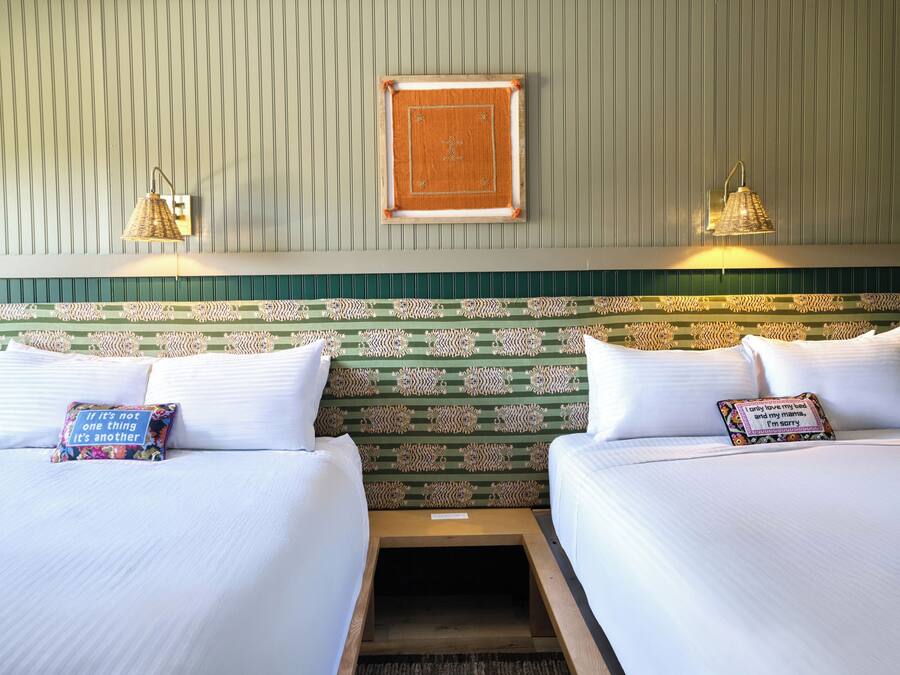 A hotel room with two beds, a patterned headboard, wall-mounted lamps, and decorative pillows.