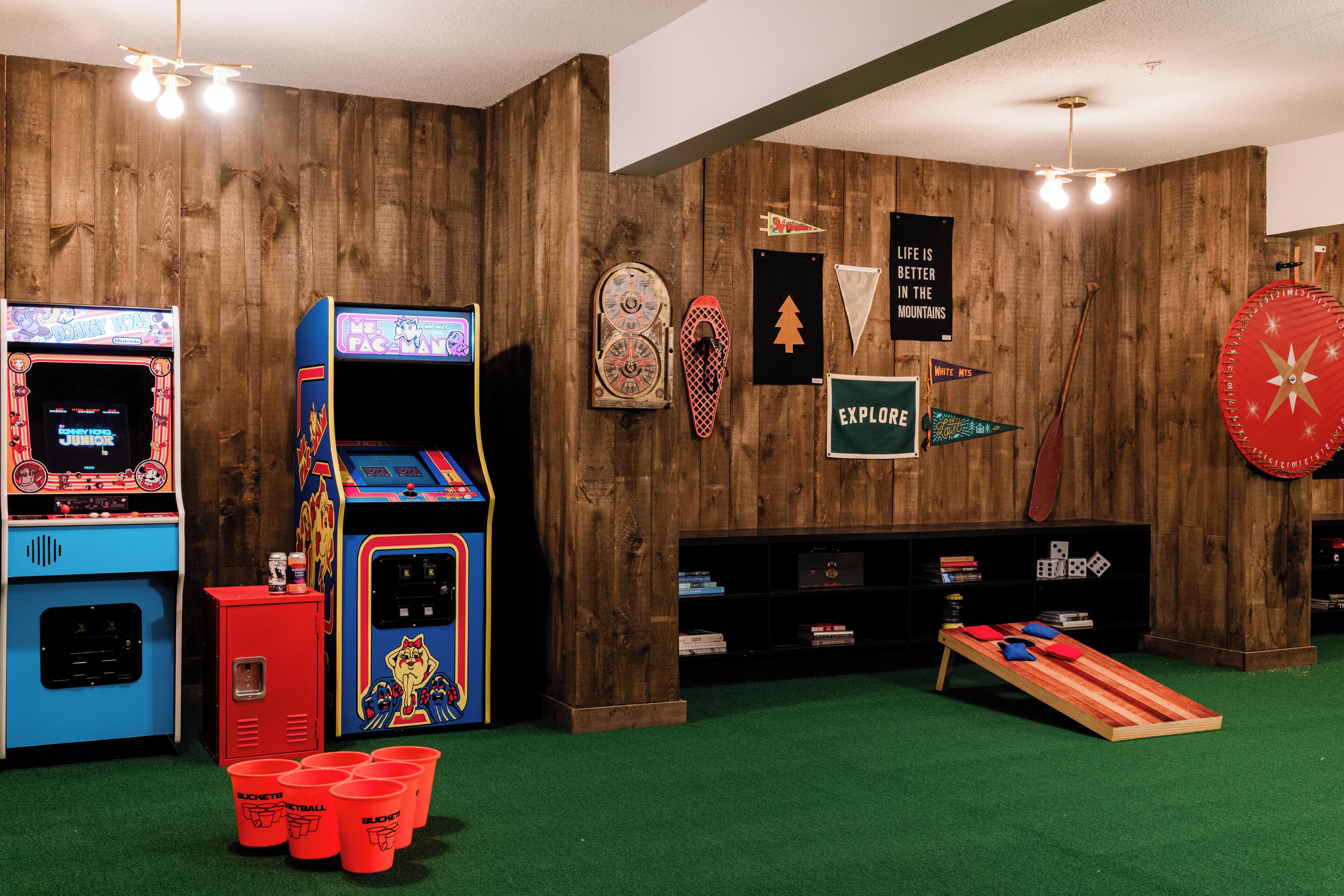 A room with wooden walls, arcade games, a dartboard, and a small table with cushions.