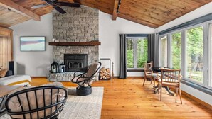A cozy room with a stone fireplace, wooden ceiling beams, and large windows. - [Goldenwood] Luxury 5 BR Winter Retreat, Fireplace (Huntsville)