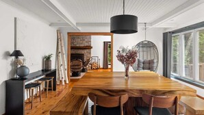 A modern dining area with a wooden table, chairs, a hanging light fixture, and a view through a large window. - [Goldenwood] Luxury 5 BR Winter Retreat, Fireplace (Huntsville)