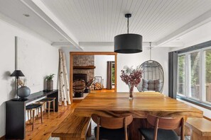 A modern dining area with a wooden table, chairs, a hanging light fixture, and a view through a large window.