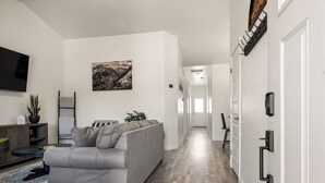 Un salon moderne avec un canapé gris, un téléviseur à écran plat, un tableau et un couloir menant à une porte.