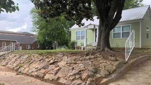 A stone retaining wall with a tree in front of a green house with a metal fence. - Cottage Studio Suite # 1 (Fort Payne)