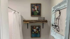 A small bathroom with a toilet, a shower curtain, a trash bin, and two wall-mounted shelves with decorative items. - Cottage Studio Suite # 1 (Fort Payne)