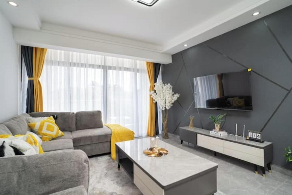 A modern living room with a grey sofa, yellow cushions, a coffee table, and a TV mounted on a dark wall.