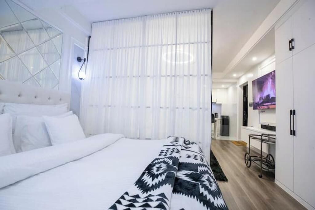 A modern hotel room with a large bed, a white headboard, a patterned throw blanket, a window with sheer curtains, a wall-mounted TV, and a kitchenette area.