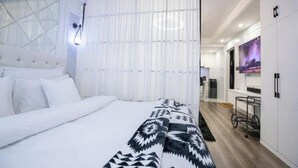 A modern hotel room with a large bed, a white headboard, a patterned throw blanket, a window with sheer curtains, a wall-mounted TV, and a kitchenette area.