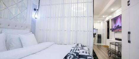 A modern hotel room with a large bed, a white headboard, a patterned throw blanket, a window with sheer curtains, a wall-mounted TV, and a kitchenette area.