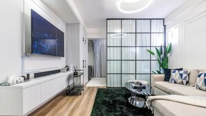 A modern living room with a large flat-screen TV mounted on the wall, a white sofa with patterned cushions, a coffee table, and a green plant.
