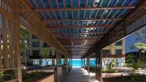 A covered walkway with a wooden ceiling and glass panels, leading to a building with a view of the sea.