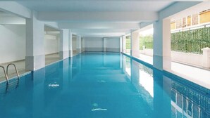 A long, narrow indoor swimming pool with clear blue water, surrounded by white walls and columns, and a view of greenery outside. - Linden Lane Hideaway Gym,Heated Pool & City Views (Nairobi)