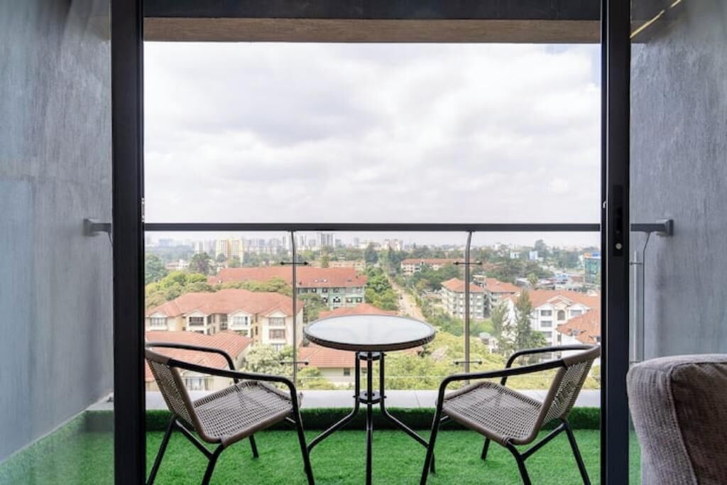 En balkon med et rundt bord og to stole, med udsigt over et boligområde med huse og bygninger.