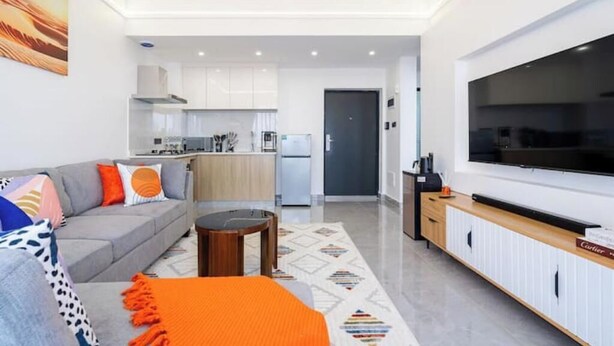 A modern living room with a grey sofa, orange throw, and a flat-screen TV. - Linden Lane Hideaway Gym,Heated Pool & City Views (Nairobi)