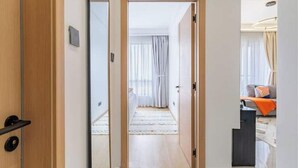A modern hotel room with a wooden door, a bed with white bedding, a small desk, and a view of the city through large windows. - Linden Lane Hideaway Gym,Heated Pool & City Views (Nairobi)