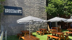 An outdoor seating area with wooden tables and striped umbrellas, a stone wall, and a sign reading "GROSSMAN'S".