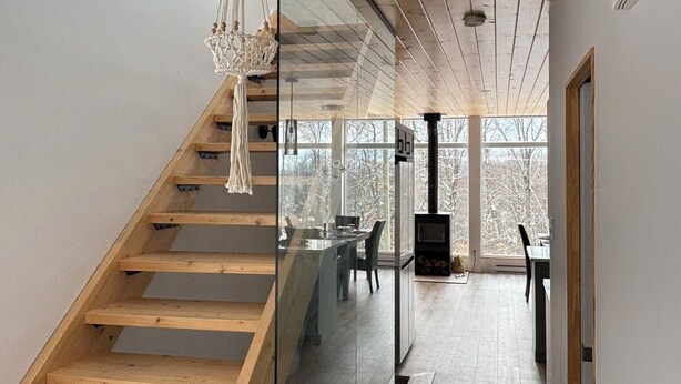 A modern interior with a wooden staircase, a glass wall, and a view of a dining area with a fireplace. - Summit Living Redefined: Alpine Views, Forest Serenity,5 Minutes from everything (Saint Donat)
