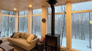 A cozy living room with a wooden ceiling, large windows, a sofa, a small table, and a wood-burning stove. - Summit Living Redefined: Alpine Views, Forest Serenity,5 Minutes from everything (Saint Donat)