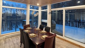 A dining area with a wooden table set for six, large windows, and a view of a snowy landscape. - Summit Living Redefined: Alpine Views, Forest Serenity,5 Minutes from everything (Saint Donat)