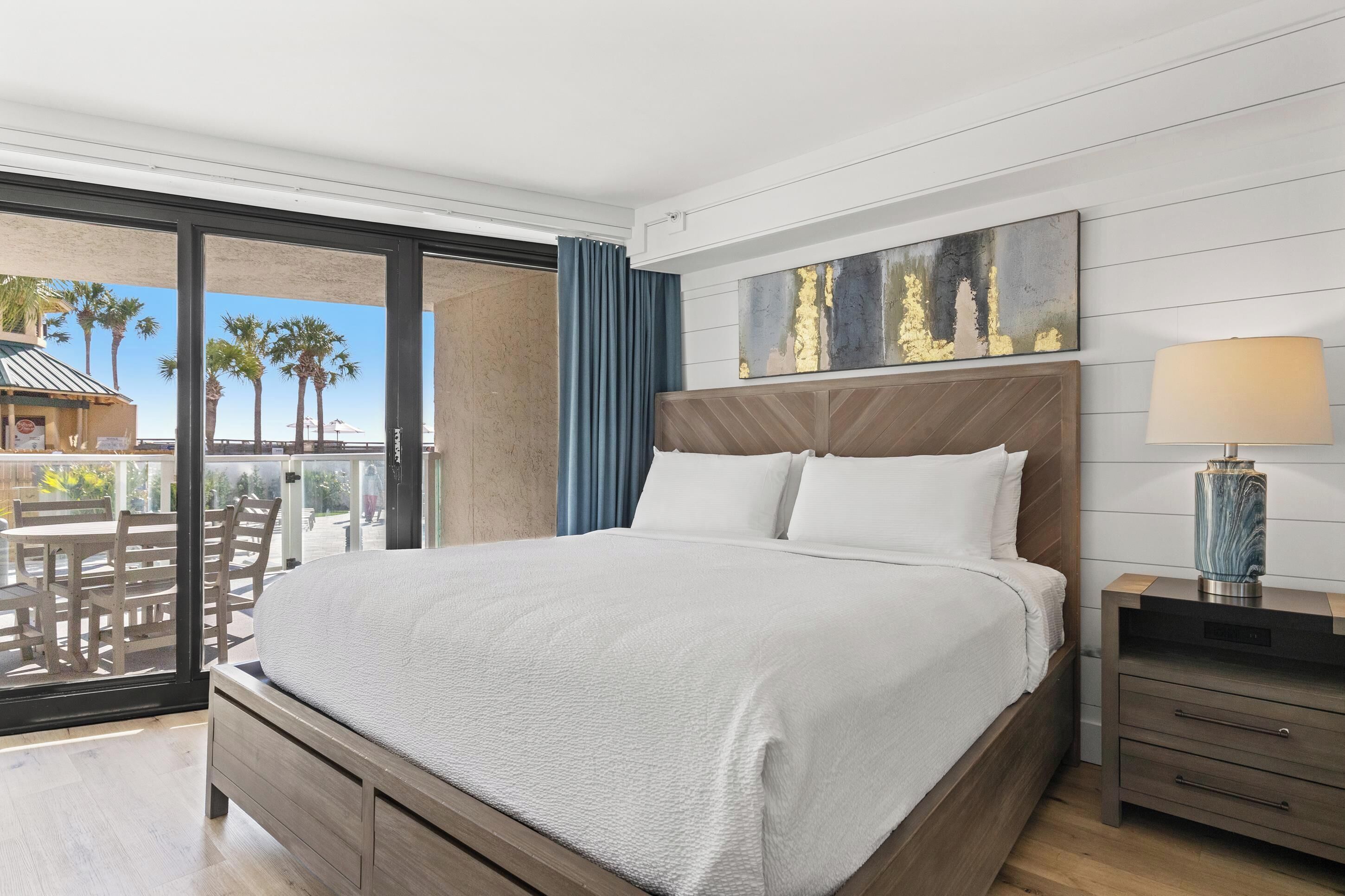 A hotel room with a large bed, a nightstand, a lamp, and a view of palm trees through a glass door.