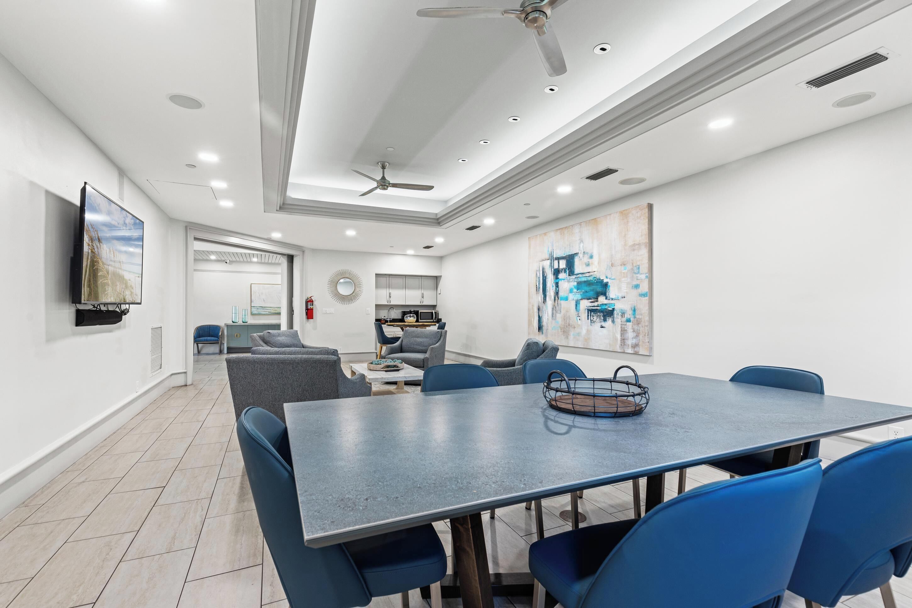 A modern dining area with a large table and blue chairs, a ceiling fan, recessed lighting, and artwork on the walls.