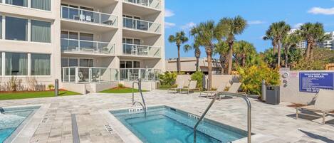 A pool area with two rectangular pools, a multi-story building with balconies, and palm trees.