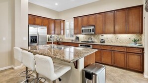 Una cocina moderna con encimeras de granito, electrodomésticos de acero inoxidable, armarios de madera y una zona de bar con taburetes blancos.