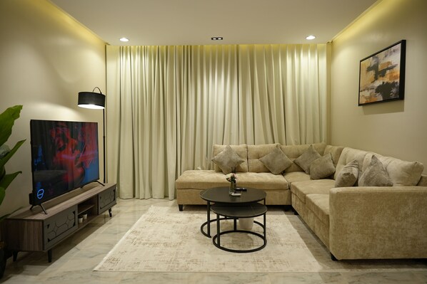 A living room with a sectional sofa, a coffee table, a TV on a stand, and a large window with curtains. - SARA Residences (Riyadh)
