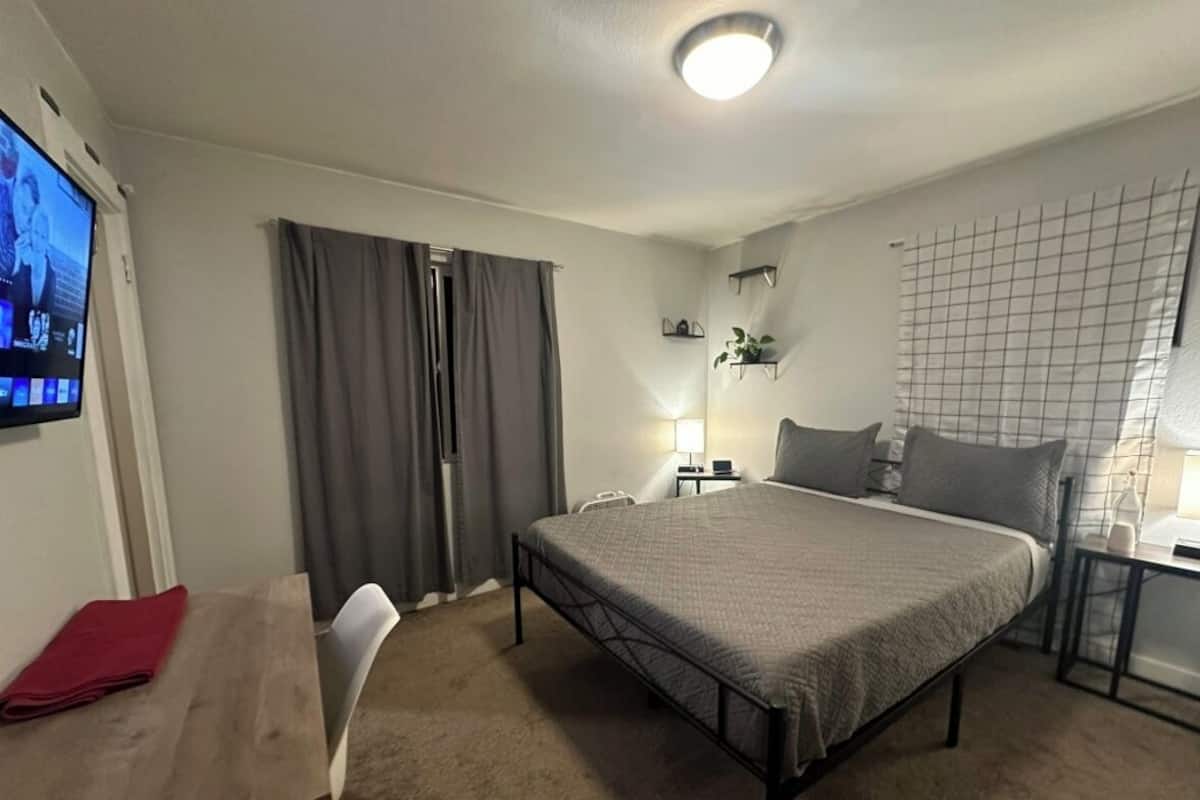 A bedroom with a bed, a desk, a chair, a TV, and a window with curtains.