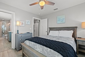A bedroom with a bed, a nightstand, a dresser, a ceiling fan, and a door leading to another room.