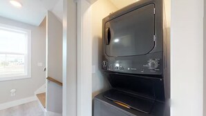 A compact laundry room with a built-in washer and dryer, a window providing natural light, and a wooden staircase leading to an upper level.