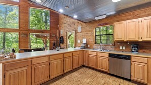 A spacious kitchen with wooden cabinets, a stainless steel dishwasher, and large windows offering views of the surrounding forest.